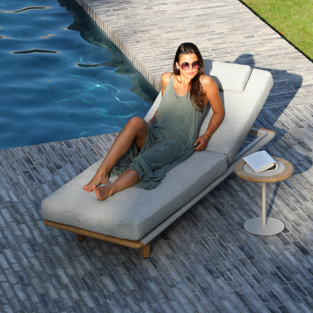 Woman relaxing beside a pool on a modern sun lounger in a luxury outdoor living space with stone paving and contemporary furniture.