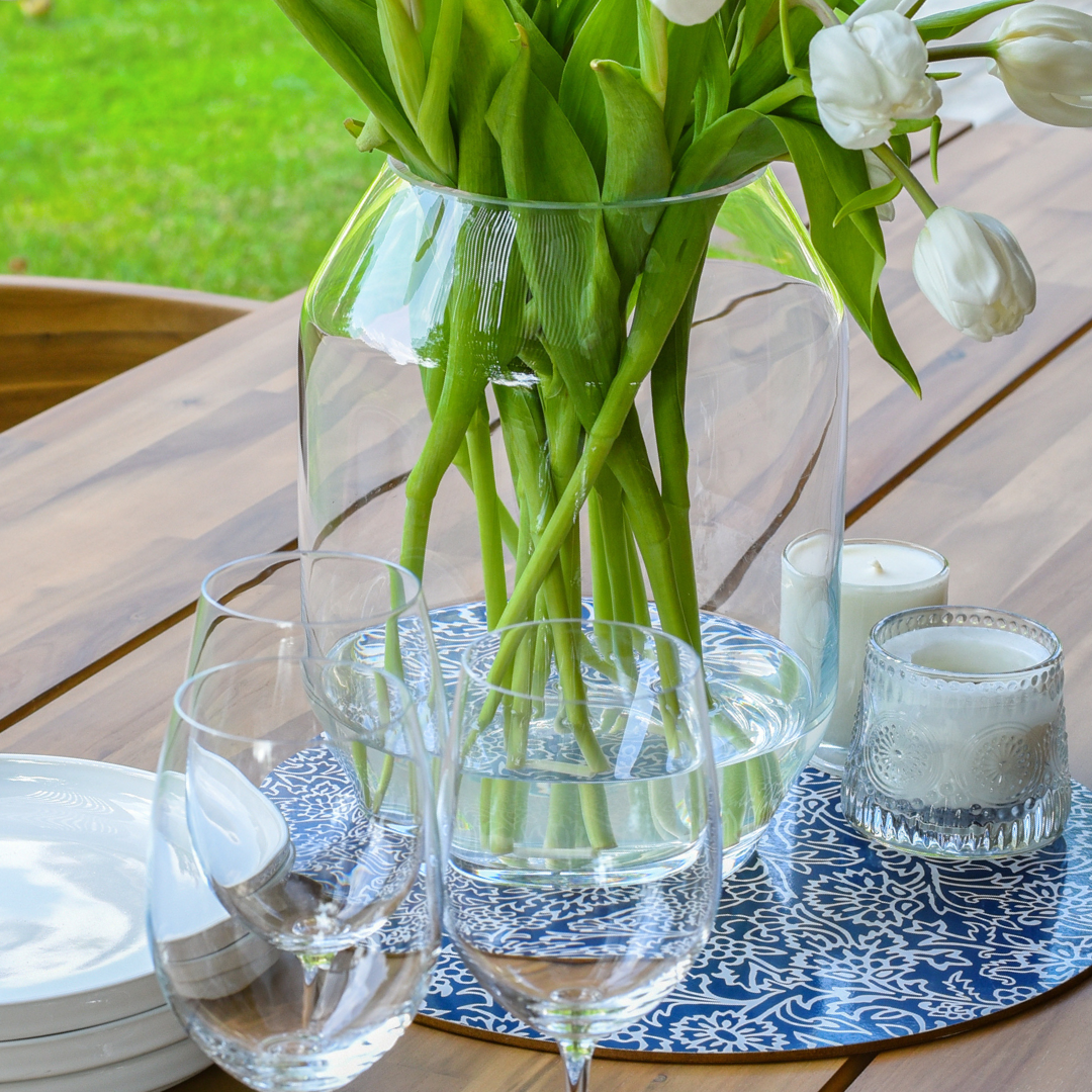 A close up image of a vase with flowers, wine glasses, candles and white plates placed on a wooden table