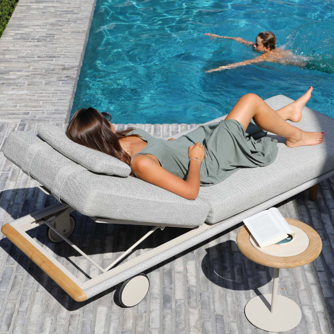 Woman relaxing on a grey upholstered poolside lounger beside clear blue water.